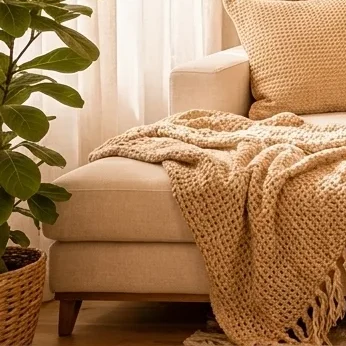 Close-up of a knitted beige throw blanket draped over a cream sofa, with a textured beige cushion and a terracotta-coloured cushion.